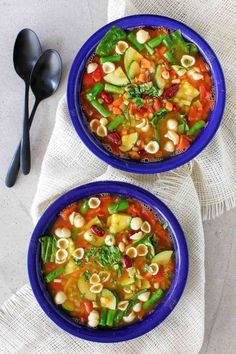 two blue bowls filled with vegetable soup on top of a white cloth next to spoons