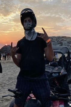 a man standing on top of a sandy beach next to a motorcycle with the sun setting in the background