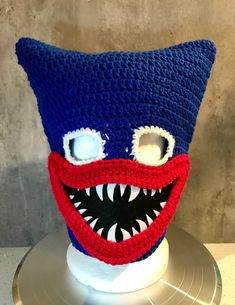 a crocheted blue hat with red and white teeth is on top of a stand