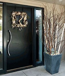 a black front door with a wreath on it and some branches in a planter