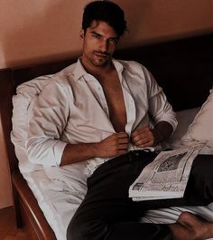 a shirtless man sitting on a bed reading a paper and looking at the camera