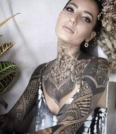 a woman with tattoos and piercings on her chest is posing for a photo in front of some plants