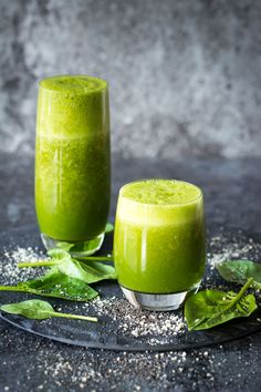 two glasses filled with green smoothie sitting on top of a black plate next to leaves