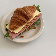 a croissant sandwich with meat, cheese and greens on a white plate sitting on a table