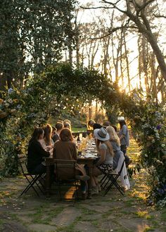 a group of people sitting around a table in the middle of a forest at sunset