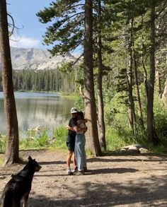 two people standing next to each other in the woods with a dog looking at them