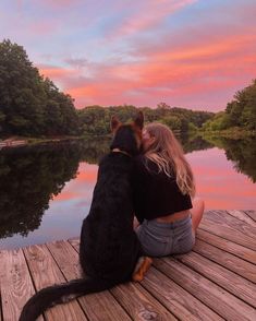 a woman and her dog are sitting on the dock looking out at the water as the sun sets