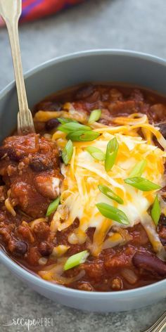 a blue bowl filled with chili and cheese on top of a gray table next to silverware