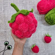 a hand holding a crocheted strawberry next to two balls of yarn