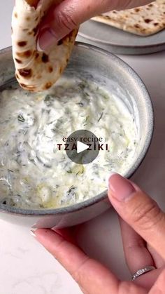 a person dipping some food into a bowl with pita bread on the side and another hand reaching for it