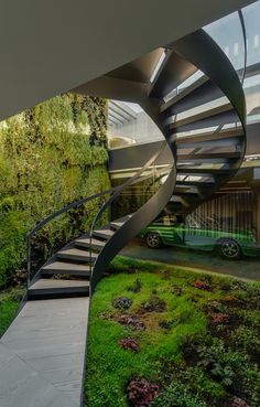 a spiral staircase in front of a house with green plants on the side and cars parked behind it