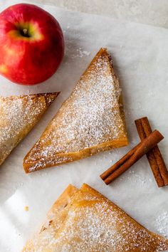 three pieces of apple pie with cinnamon sticks and an apple in the background on a piece of parchment paper