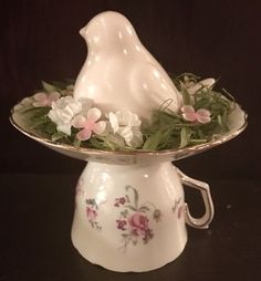 a white bird figurine sitting on top of a bowl filled with flowers and greenery