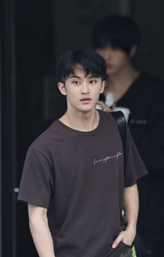 a young man in a brown t - shirt is holding a cell phone and looking at the camera