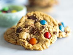 two cookies with m & m's and chocolate chips are on the table next to a bowl