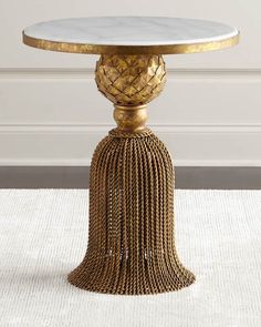 a gold and white table with tassels on the base, in front of a wall