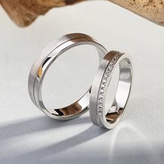 two wedding rings sitting next to each other on top of a white surface with a piece of wood in the background