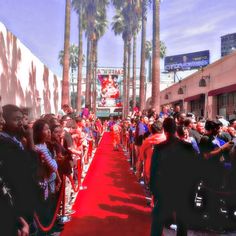 a crowd of people standing on top of a red carpet covered street next to tall palm trees