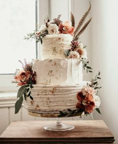 a three tiered cake with flowers and feathers on top is sitting on a table in front of a window