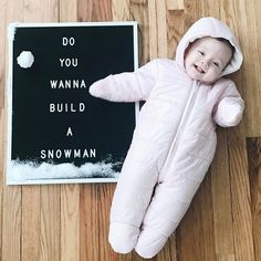 a baby in a snowsuit laying on the floor next to a sign that says, do you wanna build a snowman?