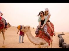 two women riding on top of a camel in the desert with other people standing around