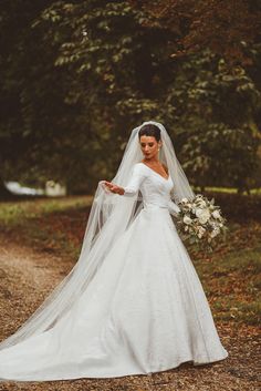 a woman in a wedding dress holding a bouquet and standing on a dirt road with trees behind her