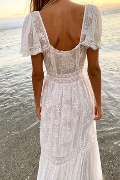 a woman in a white dress is standing on the beach looking at the water with her back to the camera