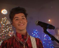 a young man standing next to a microphone in front of a christmas tree with lights