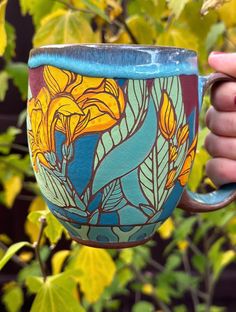 a hand holding a painted coffee cup in front of some leaves and trees with yellow flowers