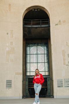 a woman in a red sweatshirt and ripped jeans poses for a photo by an arched window