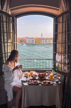 a woman sitting at a table with food and drinks in front of a window overlooking the water