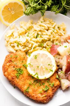 a white plate topped with meat, pasta and veggies next to lemon wedges