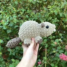 a hand holding a small stuffed animal in front of some bushes and flowers with green leaves