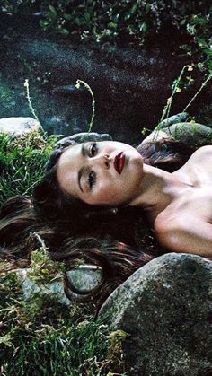 a woman laying on top of a lush green field next to rocks and plants with her eyes closed