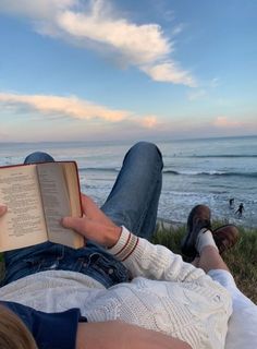 a person laying down reading a book on the grass by the beach with their feet up