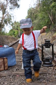 a small child wearing overalls and a hat standing next to a lantern on the ground