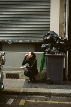 a woman sitting on the curb next to a trash can and moped parked in front of a building