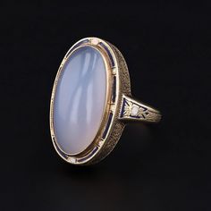 A dramatic blue-hued chalcedony ring accented with blue and white enamel. This gorgeous ring features an ethereal domed chalcedony cabochon measuring 20mm from north to south by 10mm from east to west. The chalcedony is mounted in 14k gold and the stone is framed with alternating blue and white enamel with matching blue and white enamel accents on the sides of the band. The mounting dates to the early 1900s (circa 1910-1920), while the chalcedony is a later replacement stone. The size 6 ring is Elegant Blue Oval Cabochon Moonstone Ring, Blue Art Deco Rings With Cabochon, Blue Cabochon Art Deco Rings, Blue Cabochon Rings Art Deco Style, Art Deco Blue Cabochon Rings, Oval Cabochon Enamel Ring Collectible, Formal Blue Oval Cabochon Moonstone Ring, Formal Blue Chalcedony Ring, Vintage Oval Chalcedony Jewelry