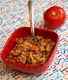 a red bowl filled with food next to a tomato on top of a colorful tablecloth