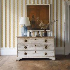 a white dresser sitting next to a painting on top of a wooden floor under a lamp