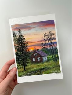 a hand holding up a small card with a painting of a cabin in the woods