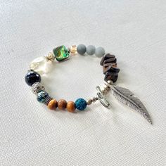 The chunky gemstone bracelet, crafted in a bohemian style, features an authentic Native American design with a silver feather toggle. This Indigenous beaded bracelet is a unique piece of jewelry, perfect for both him and her, offering a meaningful blend of tradition and style. 🪶 𝗠𝗔𝗧𝗘𝗥𝗜𝗔𝗟𝗦 + 𝗗𝗜𝗠𝗘𝗡𝗦𝗜𝗢𝗡𝗦 ❯  8mm Stones, Wood, Abalone Shell, Glass Trade Beads, Silver-plated Beads ❯  Silver-plated Toggle Clasp 🪶 𝗦𝗧𝗢𝗡𝗘 + 𝗘𝗟𝗘𝗠𝗘𝗡𝗧 𝗘𝗡𝗘𝗥𝗚𝗬 ❯  NUMEROLOGY POWER OF 3 signifying wit, charm and charisma ❯  ABALONE is known for its powers of protection ❯  CITRINE promotes creativity and intuition ❯  LAVA is a grounding stone ❯  OBSIDIAN is known to remove energetic blockages ❯  WOOD symbolizes life and growth 🪶 𝗣𝗘𝗥𝗦𝗢𝗡𝗔𝗟𝗜𝗭𝗔𝗧𝗜𝗢𝗡 + 𝗚𝗜𝗙𝗧 𝗦𝗘𝗧 𝗦𝗨𝗚? Cotton Travel Bag, Bohemian Style Jewelry, Arrow Pendant, Native American Design, Wood Necklace, Toggle Bracelet, Silver Feather, Trade Beads, Abalone Shell