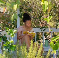 a woman sitting on a bench reading a book in the shade with trees and bushes around her