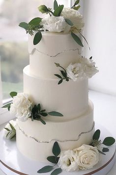 a three tiered cake with white flowers and greenery on the top is displayed in front of a window