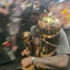 a man holding two golden trophies in front of a large group of people at a basketball game