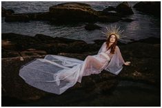 a woman in a white dress sitting on rocks near the ocean wearing a crown of jesus's head