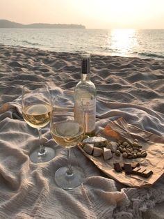 two glasses of wine are sitting on the sand at the beach with rocks and cheese