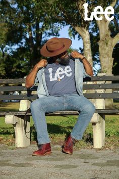 Man outdoors sitting on a park bench while posting for the camera. He is wearing brown boots, light wash straight leg denim jeans, a blue Lee logo tee t-shirt, a light wash denim button up shirt and a brown fedora hat. He is holding the collar of his open button down shirt as he is looking over his left shoulder into the distance. Spring Fashion For Men, Spring Fashion 2022, Jeans Spring Outfit, Men's Casual Outfits, Brown Fedora, Jeans Outfit Spring, Spring Outfit Idea, Light Wash Denim Jeans