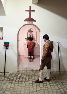 two people standing in front of a small church with a cross on it's wall