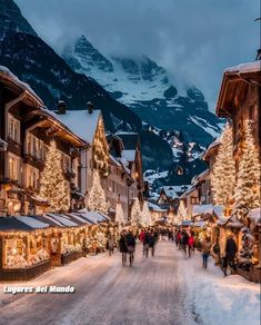 people walking down a snowy street with christmas lights on the buildings and mountains in the background